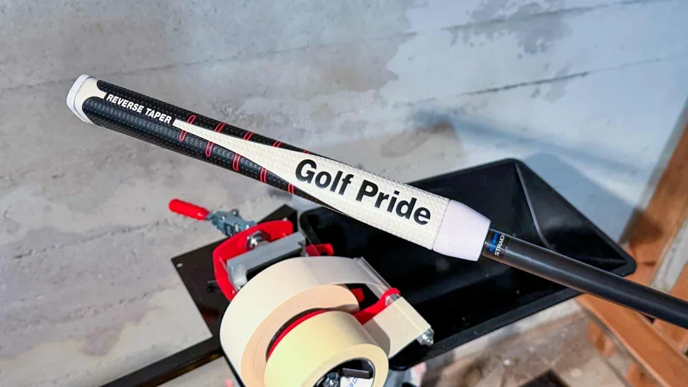 A close-up of a golf club with a white Golf Pride grip and Reverse Taper label, resting on a workbench next to a tape dispenser in a workshop setting.